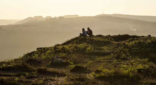 This landscape photograph shows a couple sitting on Curbar Edge, which is a prominent gritstone escarpment in the Peak District, Derbyshire, United Kingdom. The scene captures Curbar Edge during a spring evening, with golden sunlight illuminating the rural landscape and casting long shadows over the greenery and rock formations. In the distance, rolling hills and patchwork fields characteristic of the Peak District extend toward the horizon, showcasing the natural beauty of the area. The photograph highlights the tranquil nature of the Derbyshire countryside, with layers of hills and scattered trees under a warm, soft light typical of spring evenings.
